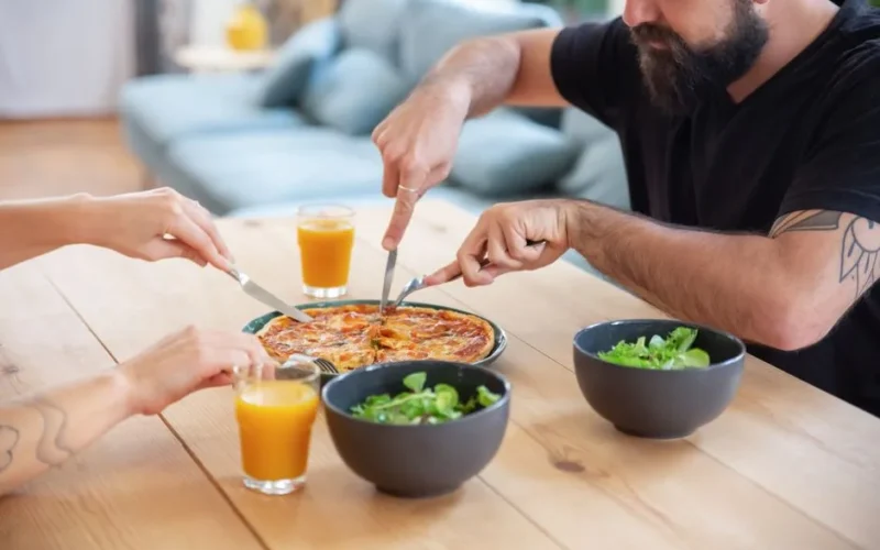 Three people enjoying pizza with orange juice and salad at a wooden table indoors.