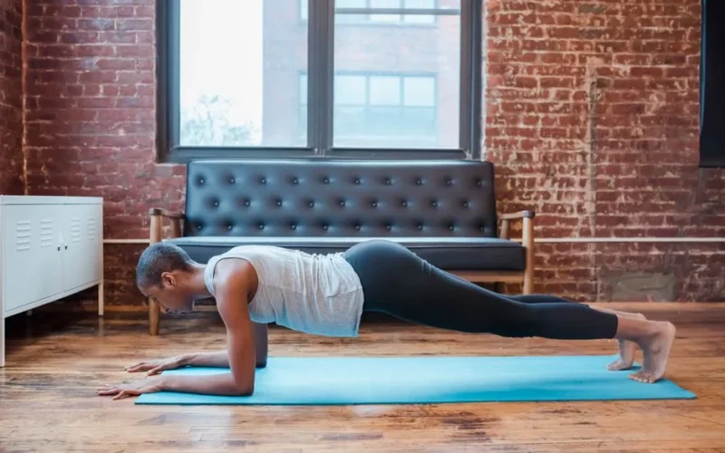 Full body side view of barefoot sportive African American female practicing Phalakasana posture on mat during yoga training at home