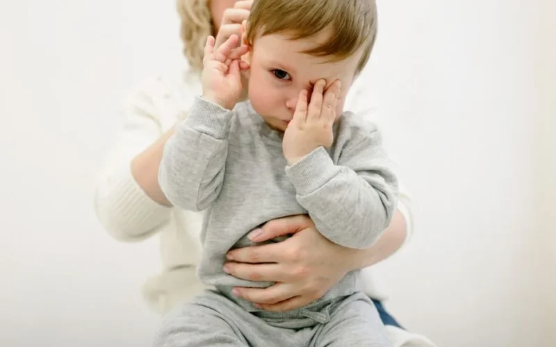 Cute baby in grey, held by adult, covering eyes playfully. Warm and cozy indoor setting.