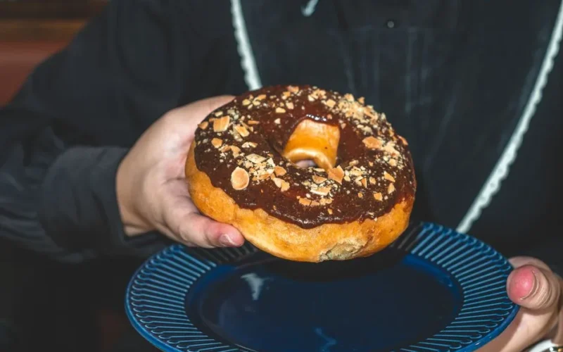 Close-up of a hand holding a chocolate donut topped with nuts on a blue plate.