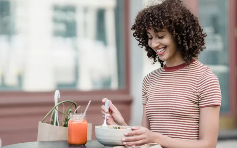 Cheerful young female with bowl of yummy food and jar of refreshing beverage sitting at table in urban cafeteria