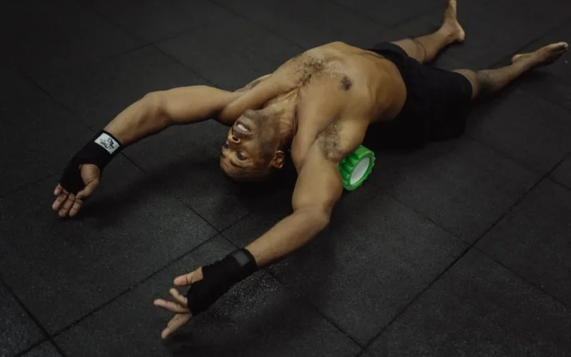 Topless man using foam roller for muscle recovery in gym setting.