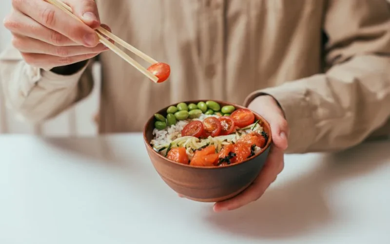 A person holding a rice bowl with cherry tomatoes, edamame, and more, using chopsticks.