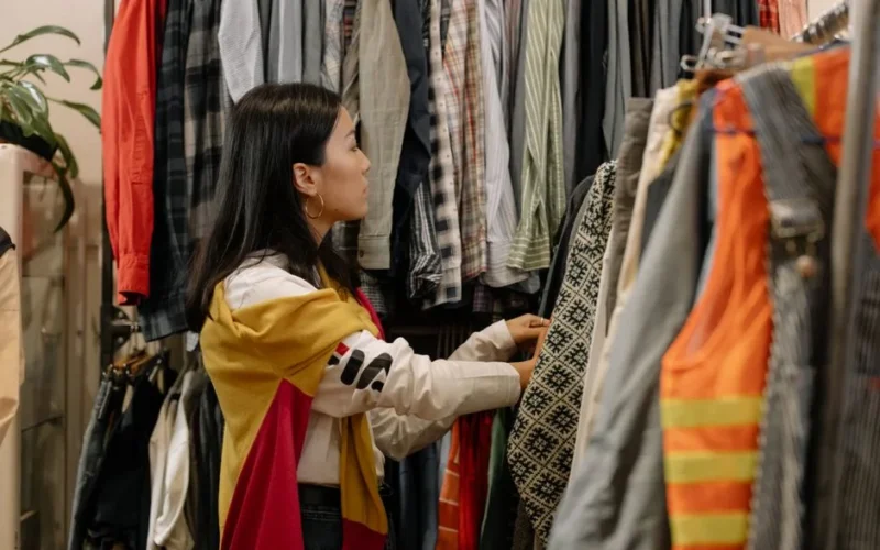 A young woman browsing clothes in a vintage store with colorful outfits.