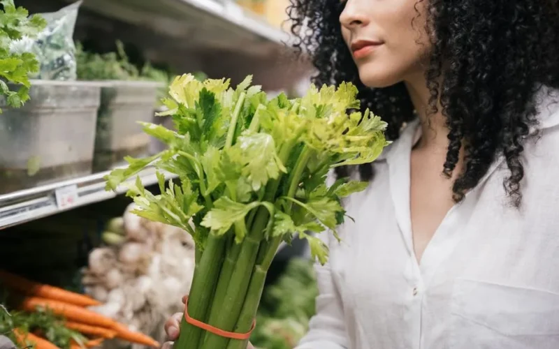 A woman examines a fresh bunch of celery in a produce aisle at the grocery store.
