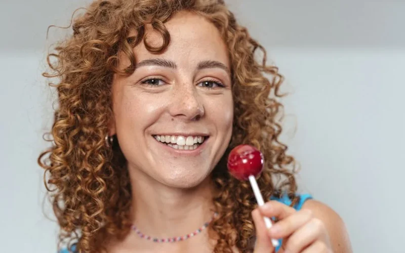 Happy woman with curly hair holding a red lollipop, smiling warmly.