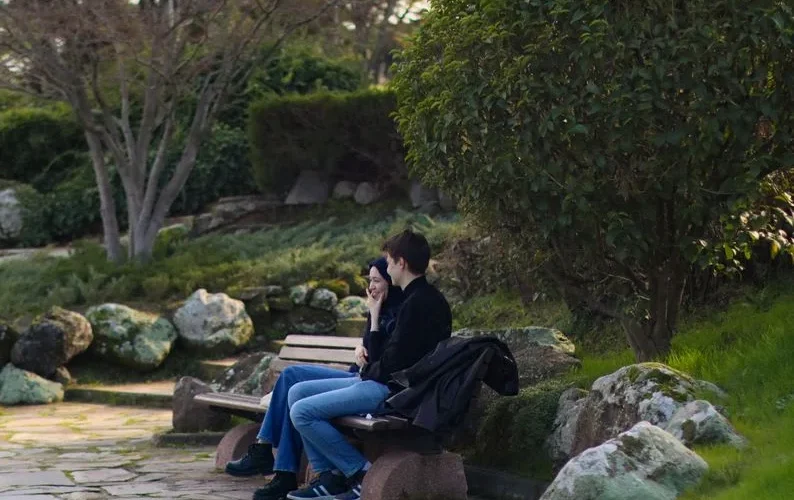 Two people sitting on a park bench surrounded by lush greenery, enjoying a quiet moment.