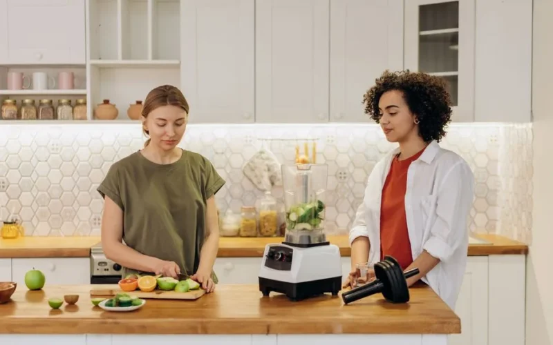 Two women preparing a healthy meal together in a modern kitchen, using fresh vegetables and a blender.