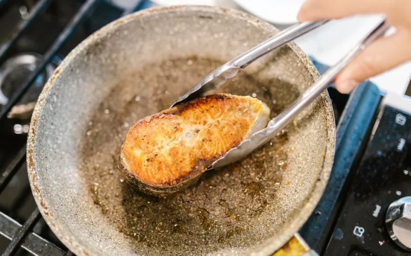 Close-up of salmon being cooked with tongs in a hot frying pan on a stove.