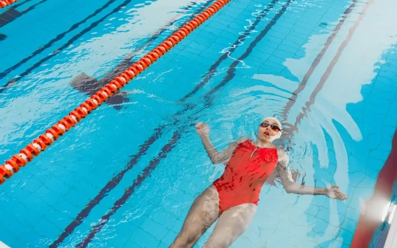 A woman in a red swimsuit floats on her back in an indoor swimming pool, enjoying a relaxing swim.