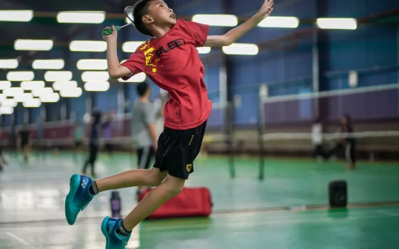 A young boy enthusiastically jumps to hit a badminton shuttlecock inside a sports hall.