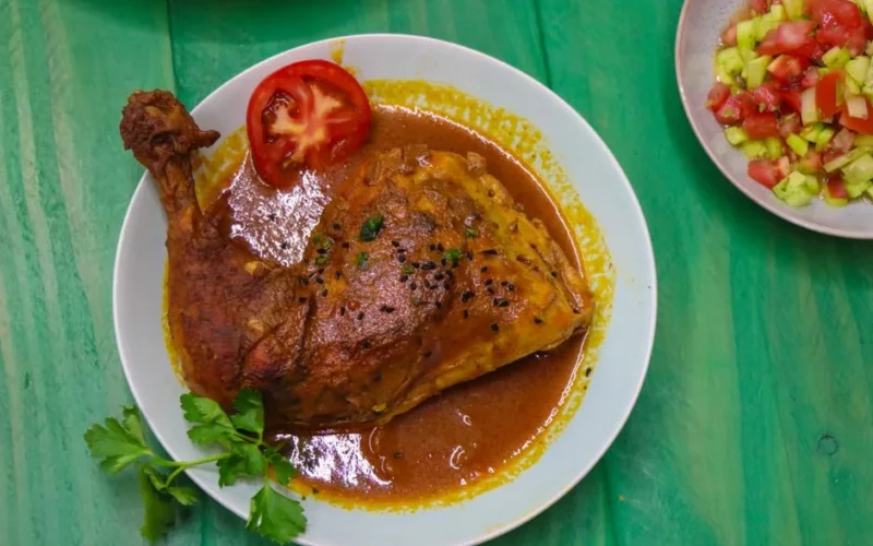 Baked chicken leg with rich sauce, garnished with fresh tomato and salad on a vibrant green table.