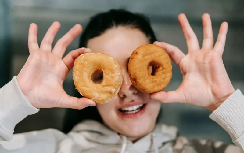 Positive young female in casual clothes smiling while covering eyes with delicious doughnuts
