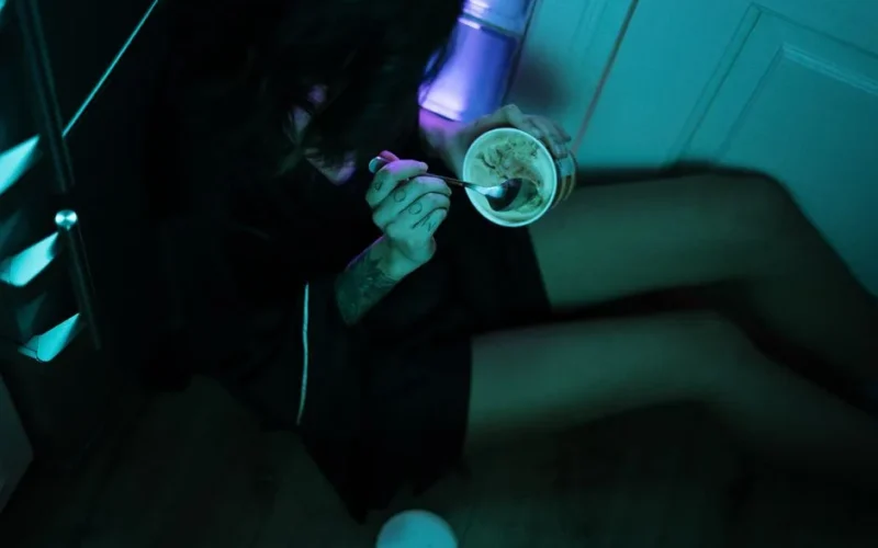 A woman enjoys ice cream on the floor of a dimly lit kitchen with neon lights, conveying isolation.