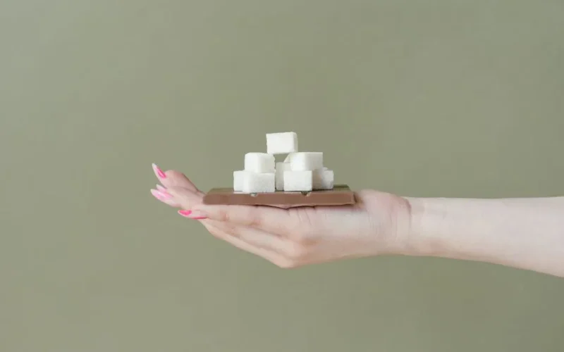 Close-up of a hand holding sugar cubes stacked on a chocolate bar against a plain background.