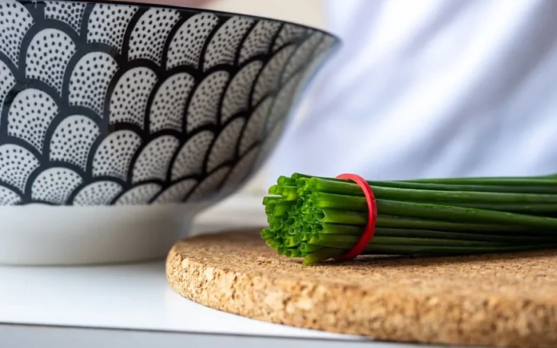 Close-up of a decorative ceramic bowl beside a fresh bunch of chives on a cork mat.