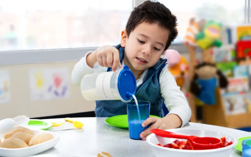 A 3-year-old pours milk into a glass while sitting at a table and smiling in a family child care home.