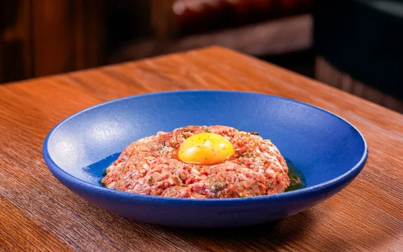 Appetizing beef tartare topped with an egg yolk served in a modern blue dish on a wooden table.