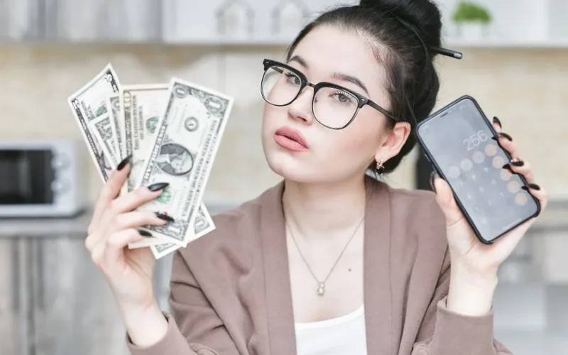 Woman holding cash and smartphone displaying calculator, highlighting personal finance management.