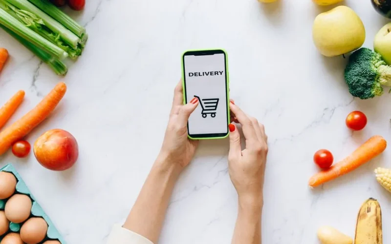 A person using a smartphone for grocery delivery surrounded by fresh fruits and vegetables.