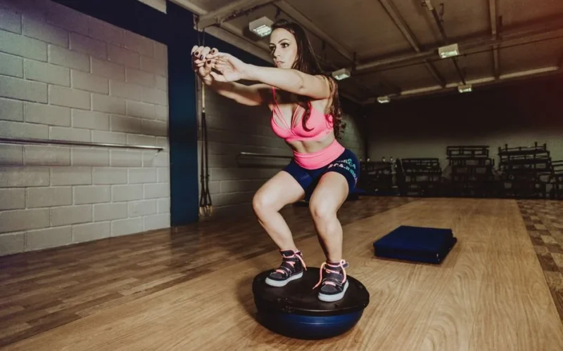Full body concentrated fit sportswoman in sports bra and shorts performing squats while balancing on bosu ball during workout in modern fitness center