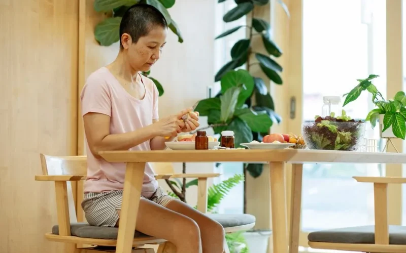 Serious adult ethnic woman with short dark hair in comfy clothes sitting at wooden table with bowl of salad and medicines while peeling healthy fruits at home