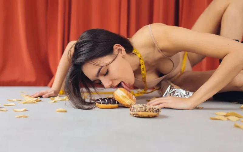 A woman in a spaghetti strap dress eating donuts on the floor, surrounded by chips, with a measuring tape around her neck.