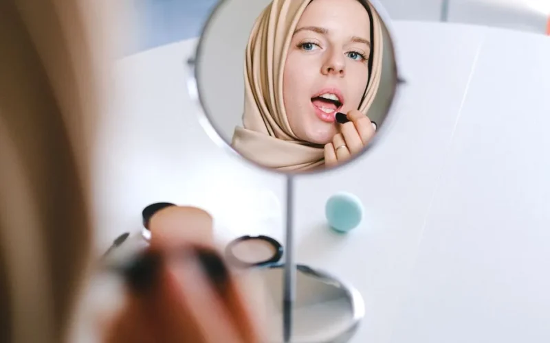 Muslim woman in hijab applies lipstick while looking at her reflection in a mirror.
