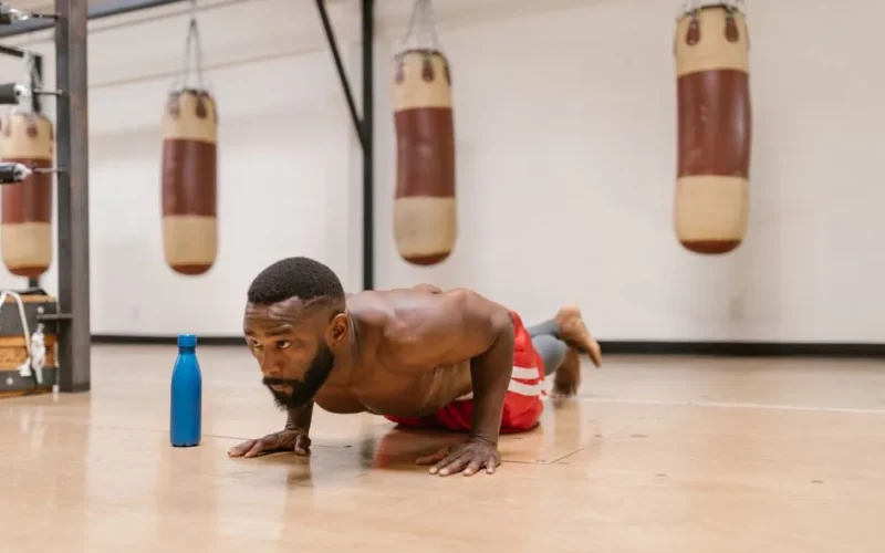 A focused shirtless man performing push-ups on a gym floor beside a blue water bottle.