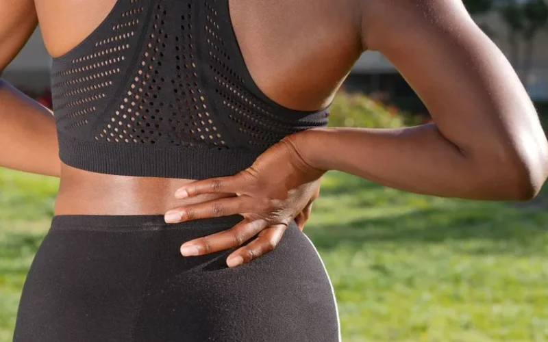 Woman in athletic attire holding her back outside, indicating discomfort or pain.