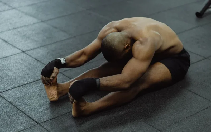 A shirtless man stretches on a gym floor, barefoot and wearing black shorts, focused on flexibility.