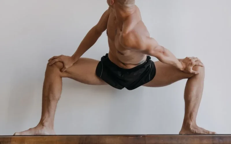 A shirtless man with a muscular build stretches on a wooden surface indoors.