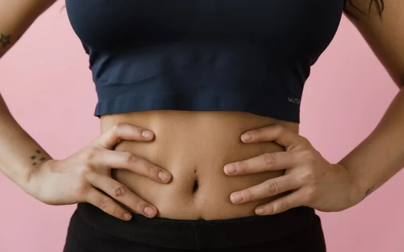A close-up shot of a woman's toned stomach with hands on hips, against a pink background.