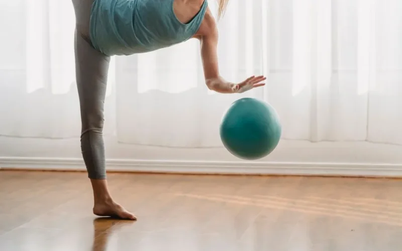 Sportive anonymous barefoot female in activewear standing on one leg while doing exercise with fitness ball during training in room