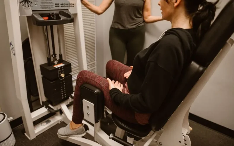 A woman performs exercises on a hip abductor machine with guidance from a trainer.