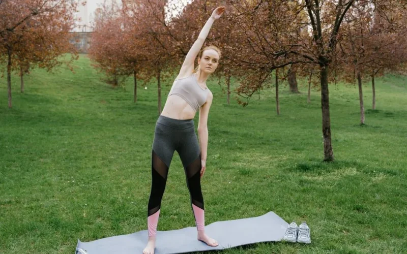Woman in activewear performing side stretch on yoga mat in a park setting
