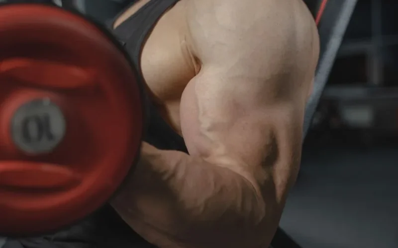 A focused view of a bodybuilder's arm muscles lifting a dumbbell in a gym setting.