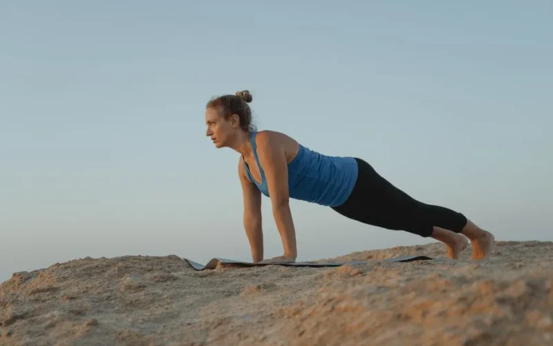 A woman performs a yoga pose on a sandy outdoor terrain under a clear sky at dusk.