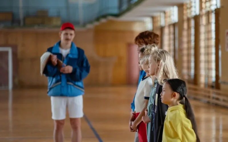 Diverse group of children standing indoors in a gym with a coach in focus.