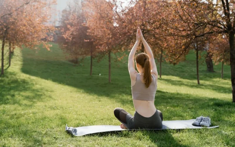 A woman practicing yoga on a mat outdoors in a sunlit park during autumn.
