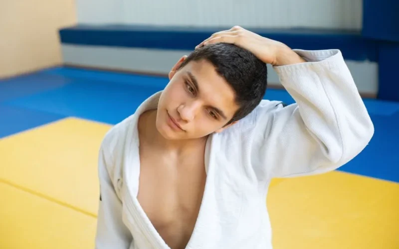 A young man in a judo uniform stretches his neck in a brightly colored gym setting, expressing focus and concentration.