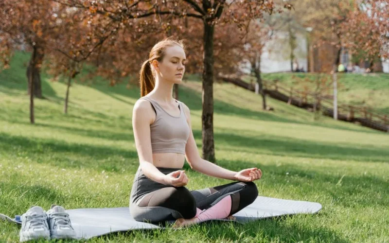 Woman meditating on a yoga mat in a park, promoting fitness and wellness.