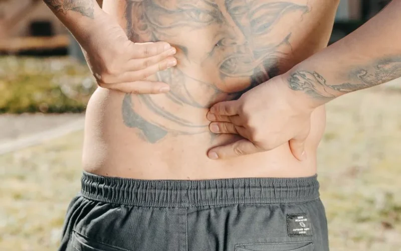 Close-up of a man holding his tattooed back outdoors, possibly experiencing back pain.