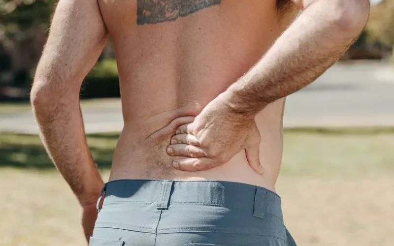 Shirtless man holding his lower back outdoors indicating pain or discomfort.