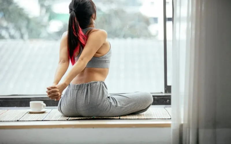 Back view of slim unrecognizable female sitting on mat and stretching arms behind back while doing warm up exercise in morning on terrace