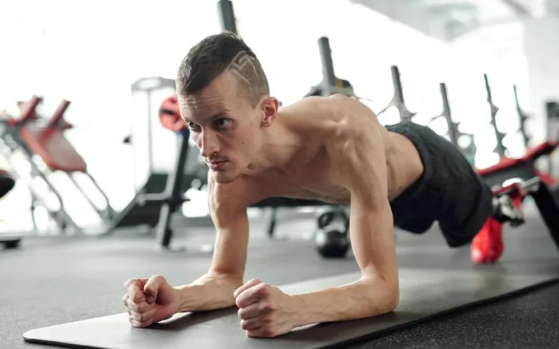 A shirtless man with a prosthetic leg planks on a yoga mat in a gym, showcasing fitness and determination.