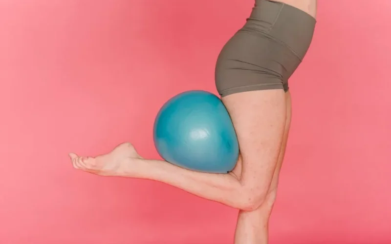 Side view of crop anonymous female stretching tiptoes while balancing on leg with fitness ball against pink background
