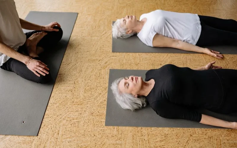 Two senior women practicing relaxation on yoga mats indoors, supervised by an instructor.