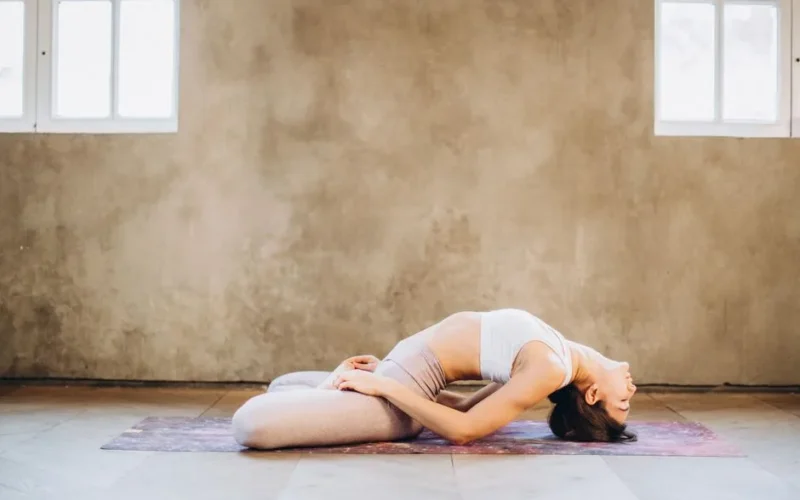 A woman performing Matsyasana yoga pose on a mat indoors, focused on relaxation and wellness.