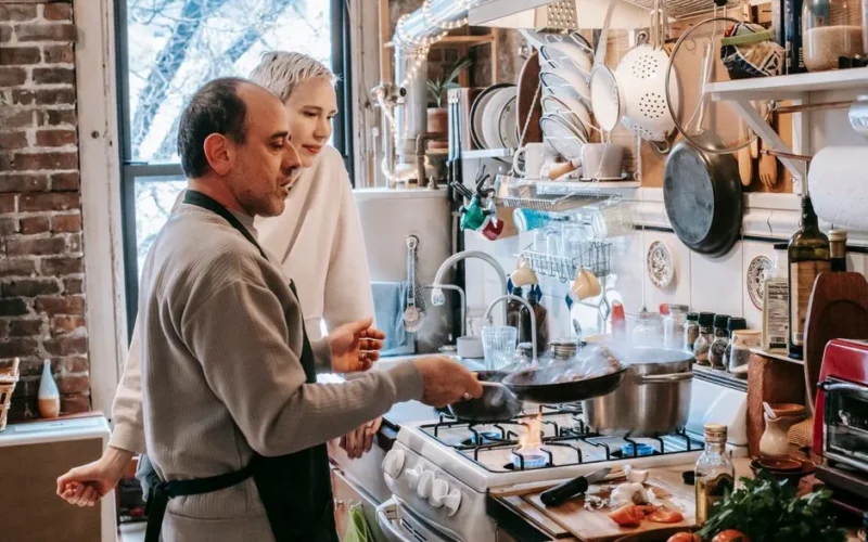 Concentrated husband in apron frying food on pan while cooking on gas stove with wife in kitchen